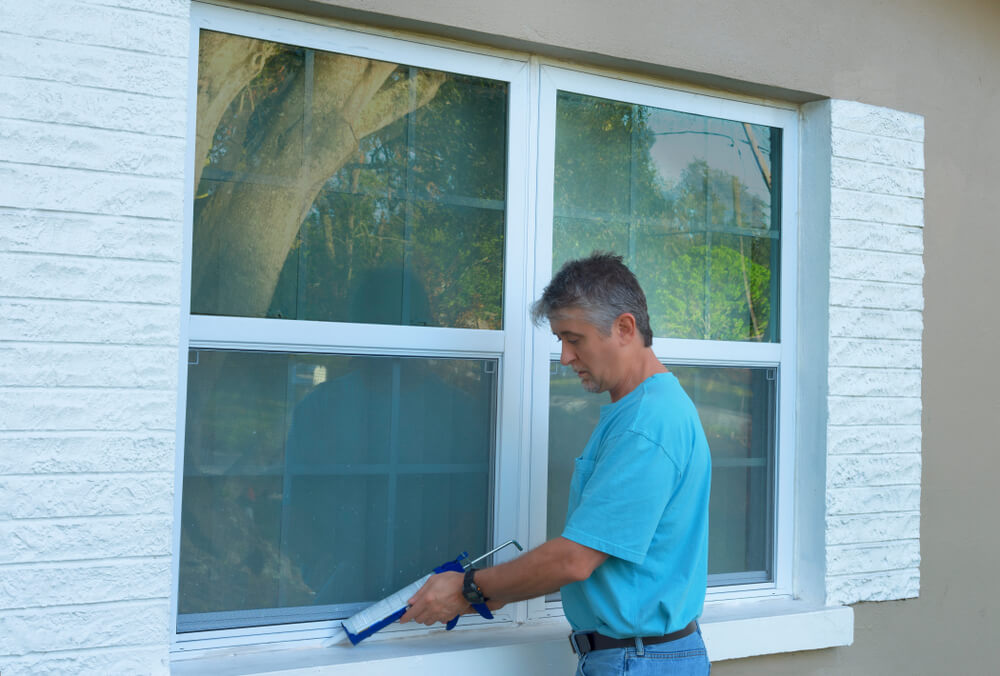 A man caulking window with a caulk gun, an important part of weatherproofing homes and houses against hurricanes.